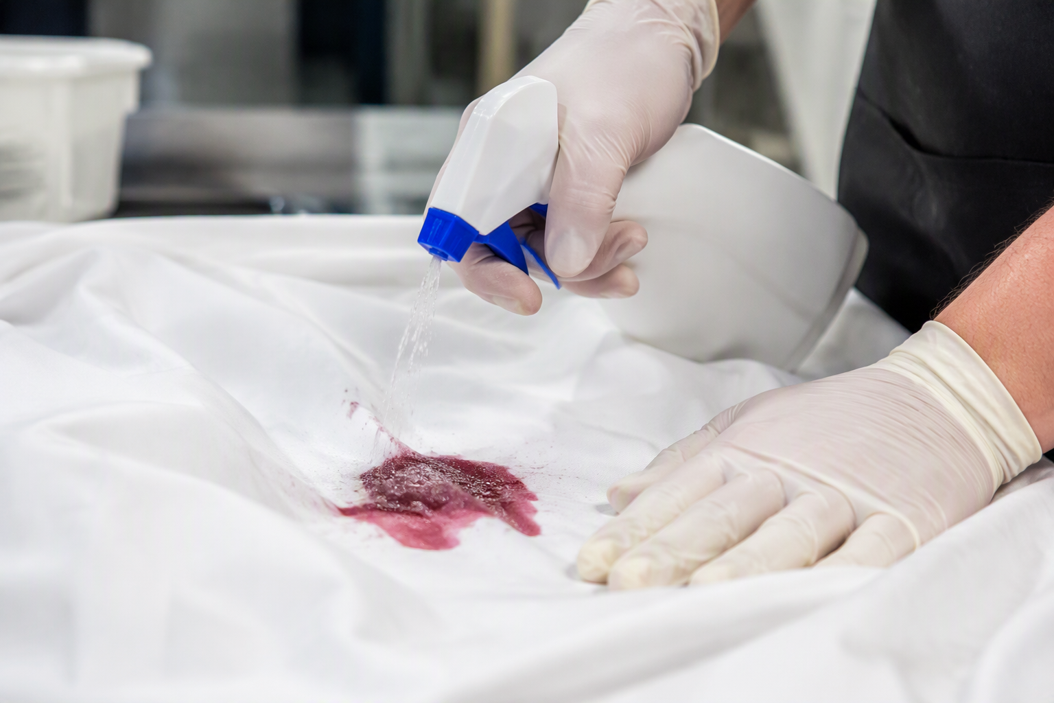 Hand applying stain remover from a spray bottle onto a red wine stain on a white tablecloth